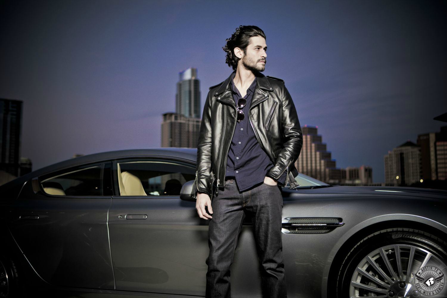 male hispanic dark haired model with 5 o'clock shadow and wearing a black leather coat and blue jeans in front of an aston martin with the austin skyline in the background at dusk